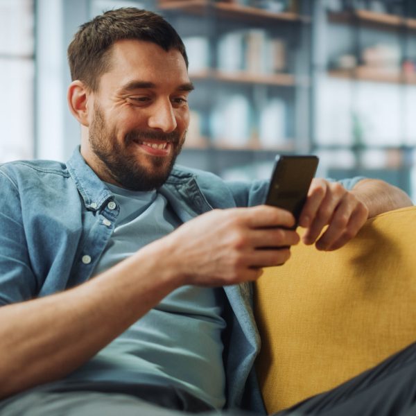 Happy Handsome Caucasian Man Using Smartphone in Cozy Living Room at Home. Man Resting on Comfortable Sofa. He's Browsing the Internet and Checking Videos on Social Networks and Having Fun.